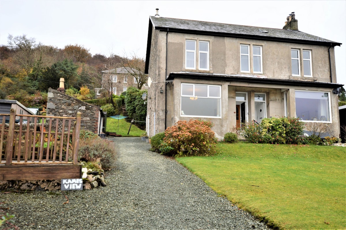 The exterior of the two-story apartment features a warm stone facade with large windows that invite natural light. A garden pathway leads to the entrance, surrounded by well-tended greenery and seasonal flowers. A sign marks the property as 'Kames View,' enhancing the welcoming atmosphere.