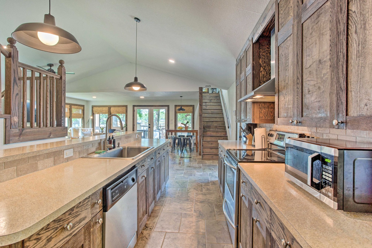 The kitchen features an open layout with wooden cabinetry and modern appliances, including a dishwasher and microwave. Natural light fills the space through large windows, while a staircase leads to the upper level. A dining table is visible in the background, complementing the inviting atmosphere.