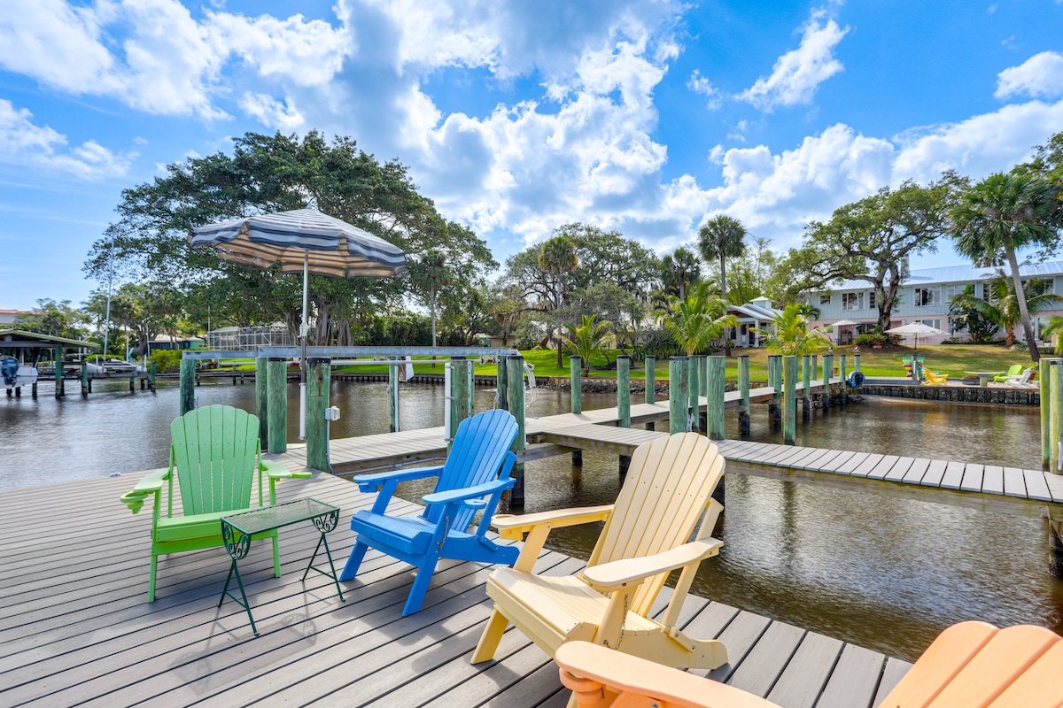 A serene dock area features colorful adirondack chairs in green, blue, and yellow, positioned for enjoying riverside views. An umbrella provides shade over a small table. The calm water reflects trees and homes in the distance, with a clear blue sky and scattered clouds overhead.