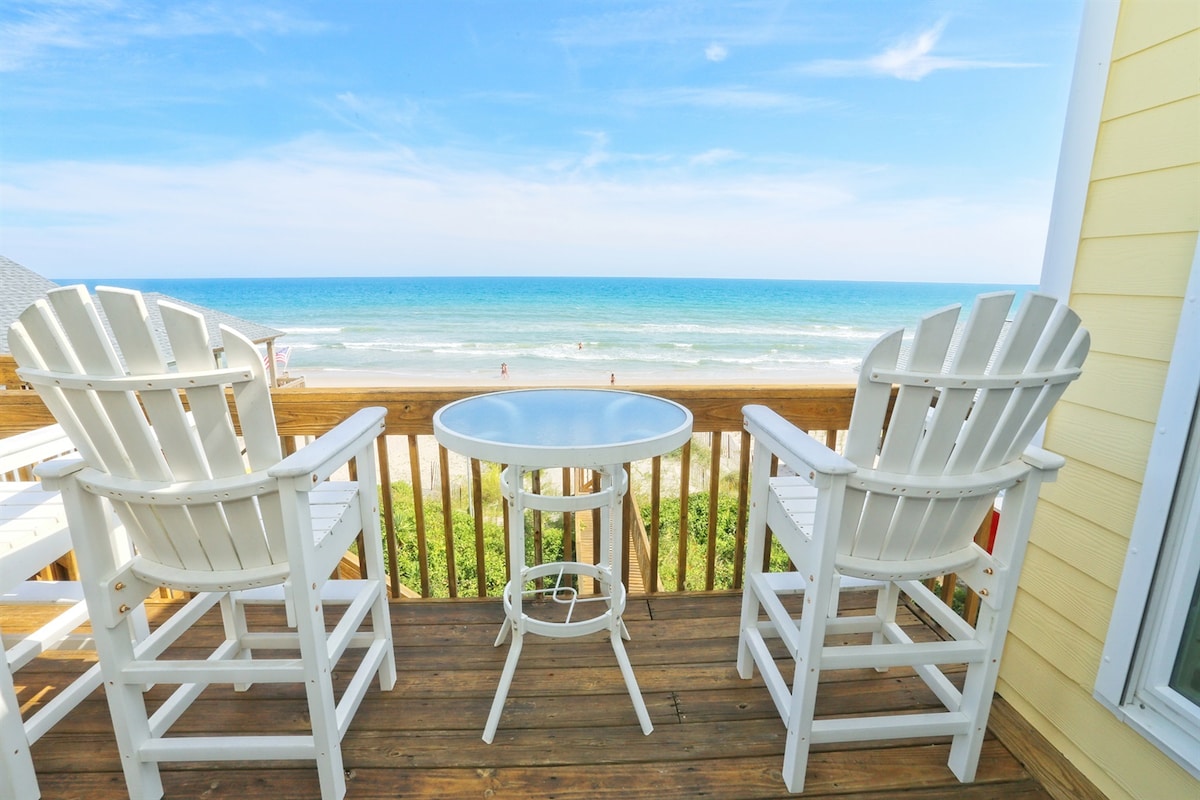 Two white Adirondack chairs flank a round table on a wooden deck, offering unobstructed views of the ocean and sandy beach. The sky is clear with gentle waves visible in the distance, creating a serene coastal setting.