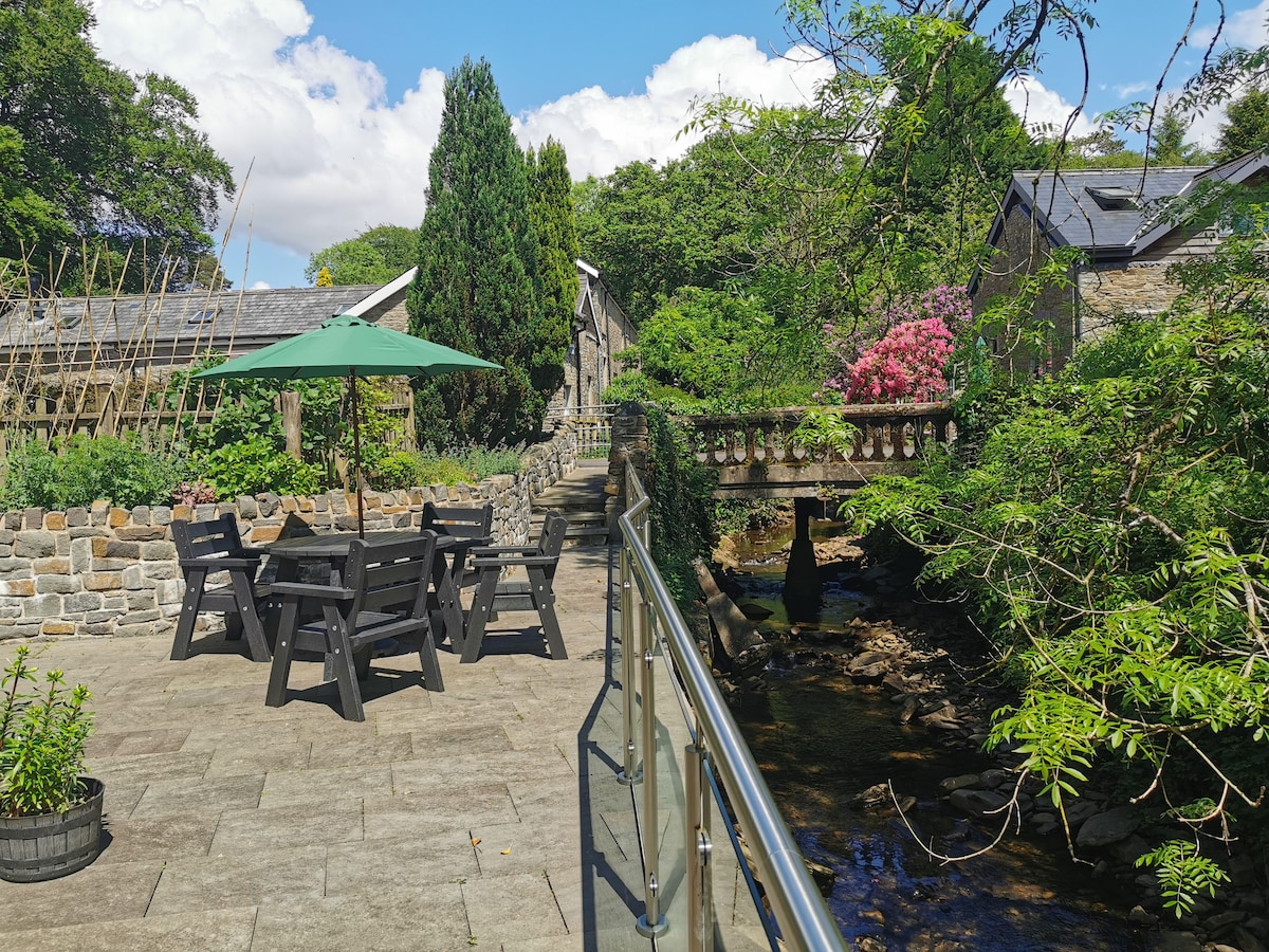 An outdoor patio area features a black wooden dining set under a green umbrella. Stone walls surround the space, and flowering plants provide vibrant color. A gentle stream flows nearby, with trees and buildings visible in the background against a clear blue sky.