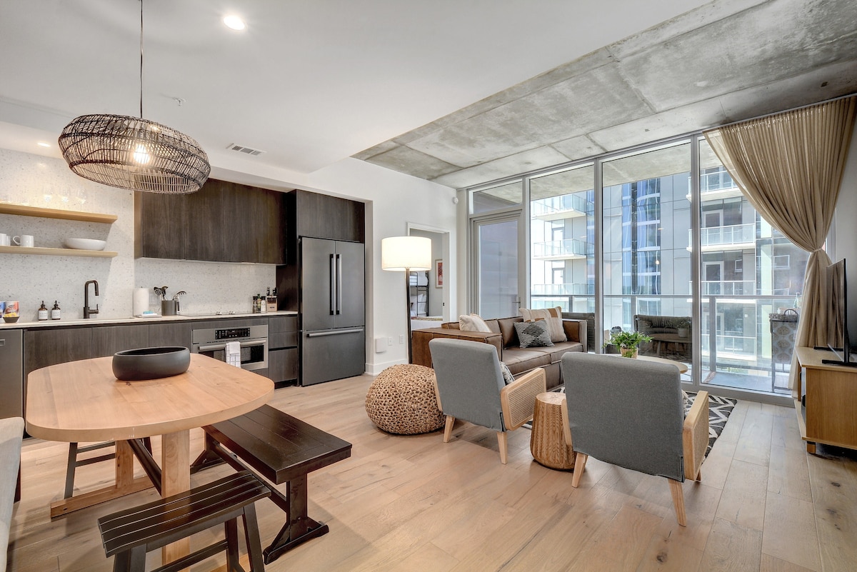 A modern open-concept living area features a round dining table with sturdy chairs beside a stylish kitchen. Comfortable seating is arranged in the lounge area, complemented by natural light flowing through large windows. Decorative elements include a woven pendant light and a neutral color palette.