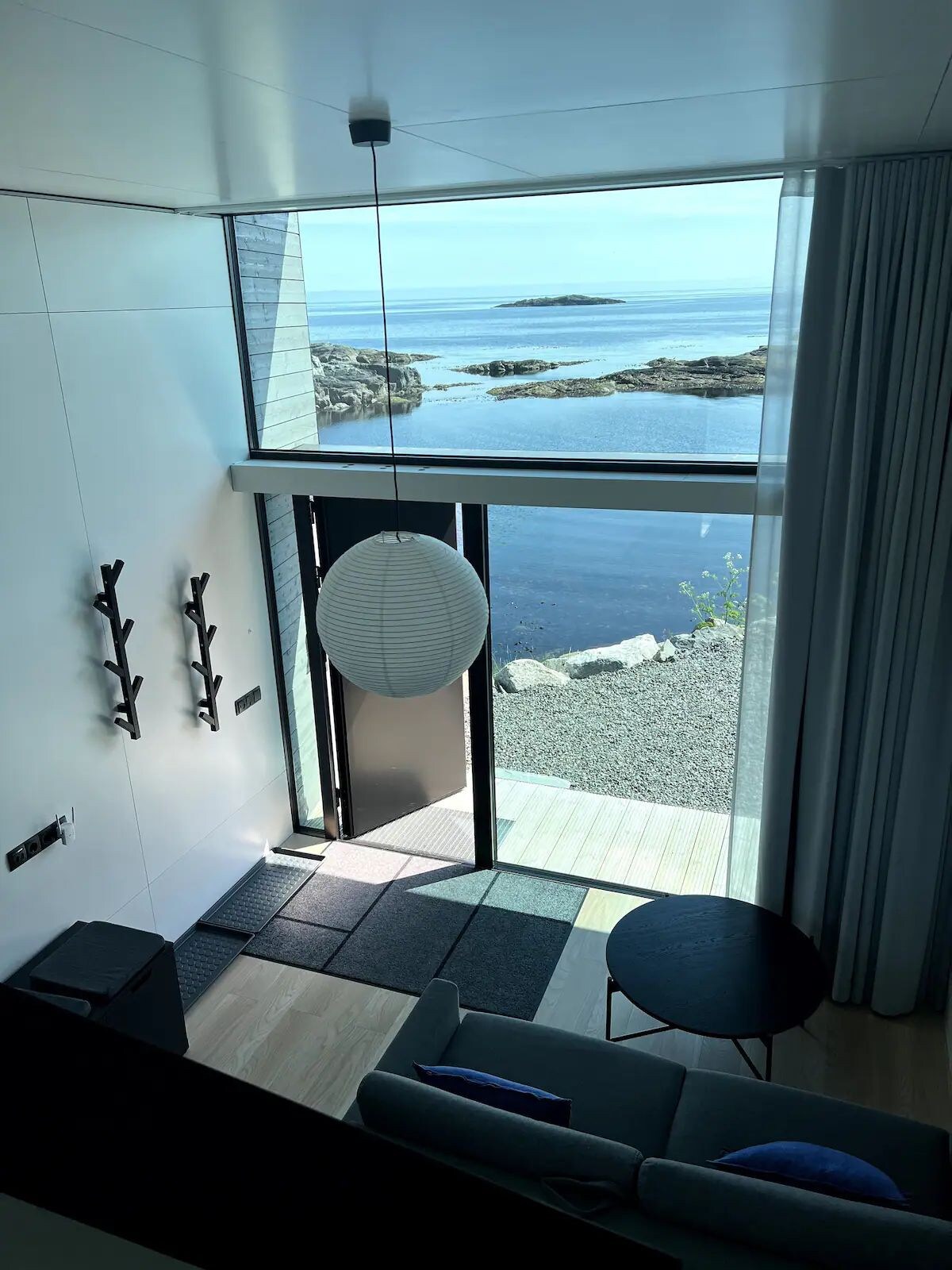 A modern entryway reveals large glass doors overlooking the water. Light grey walls and light wood flooring create a bright atmosphere. A cohesive seating area features a sofa and circular table, while a decorative pendant light hangs from the ceiling, enhancing the airy space.