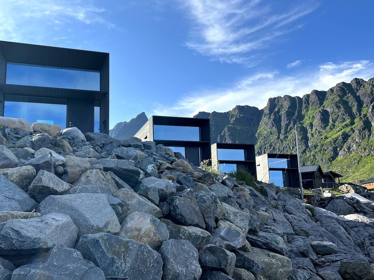 A row of modern mini homes is set against a backdrop of rugged mountains. Large glass windows reflect the sky, creating a seamless connection with the surrounding landscape. Rocky terrain leads up to the structures, enhancing the natural beauty of the site.