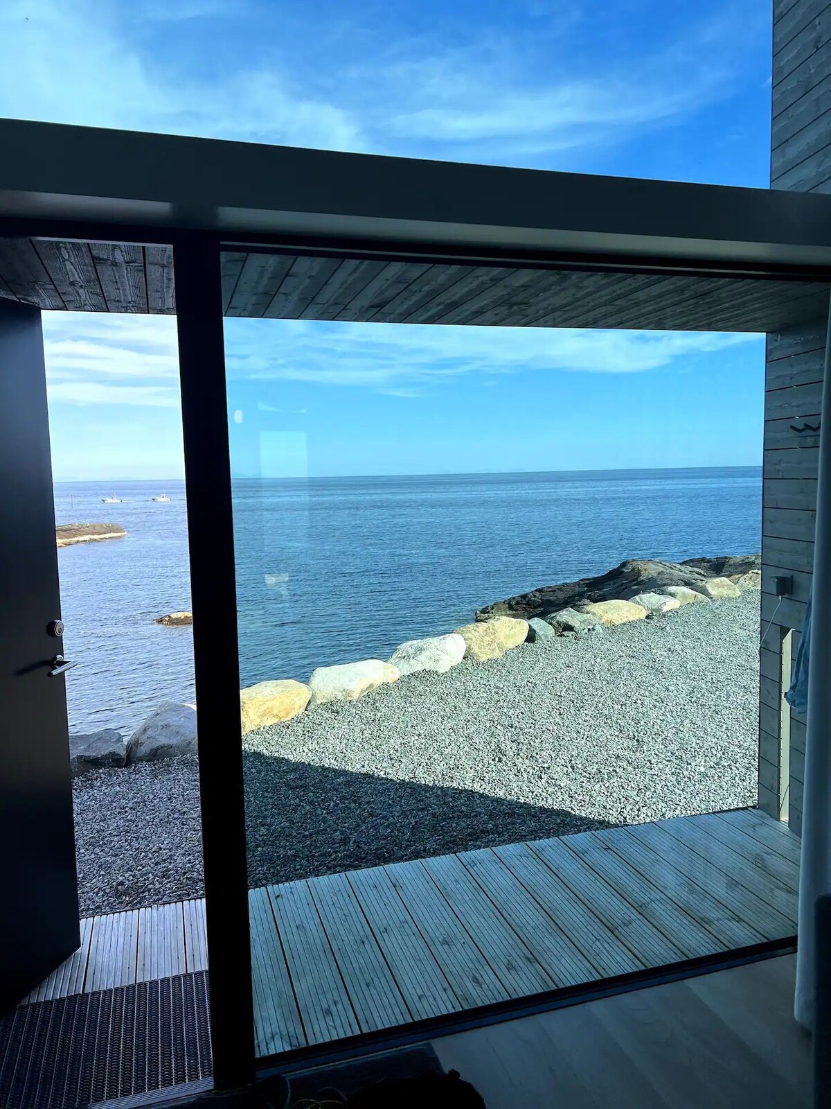 A door stands open to reveal a serene view of the ocean, framed by a modern structure. Smooth stones line the water's edge, complementing the pebbled shoreline. The sky appears bright and clear, enhancing the tranquil atmosphere outside.