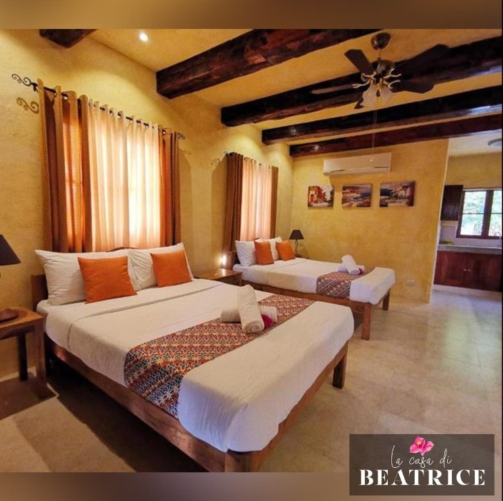 A spacious room is seen with two large beds, each adorned with simple patterned throws and complemented by neatly arranged towels. Natural light filters through large windows, illuminating the warm yellow walls and wooden beams overhead. A small lamp rests on each bedside table.