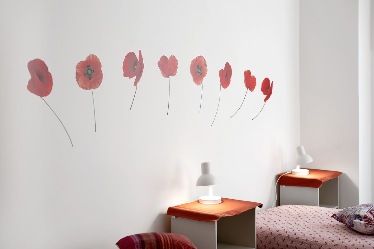 A bedroom features a soft white wall adorned with a series of red poppy flower decals, creating a cheerful accent. On either side, bedside tables are topped with minimalist lamps, while inviting bedding adds a touch of comfort to the space.