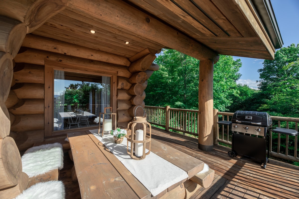A spacious outdoor area features a wooden deck with a large table and bench seating, complemented by soft, white cushions. A barbecue grill is positioned nearby, and the surrounding greenery enhances the tranquil setting, providing a serene view of the outdoors.