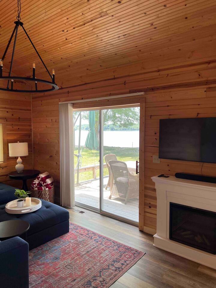 A cozy living area features warm wooden walls and a large window that opens to the lake. A dark sofa is accompanied by a side table with a lamp. A mounted TV and a fireplace are positioned along one wall, enhancing the rustic charm.