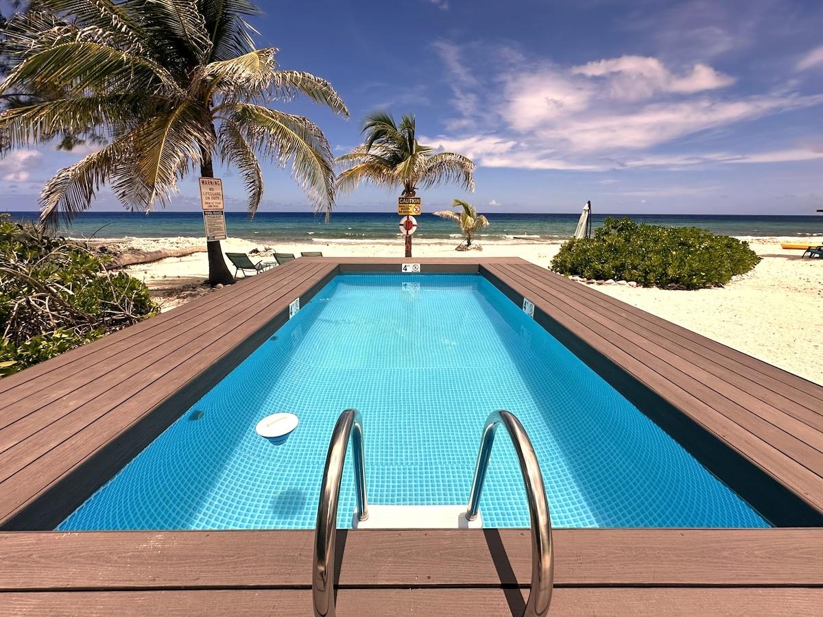 A serene pool is bordered by a wooden deck, offering clear blue water and steps leading down into it. Palm trees swaying gently in the breeze are visible, with the sea and sandy beach in the background under a bright sky.