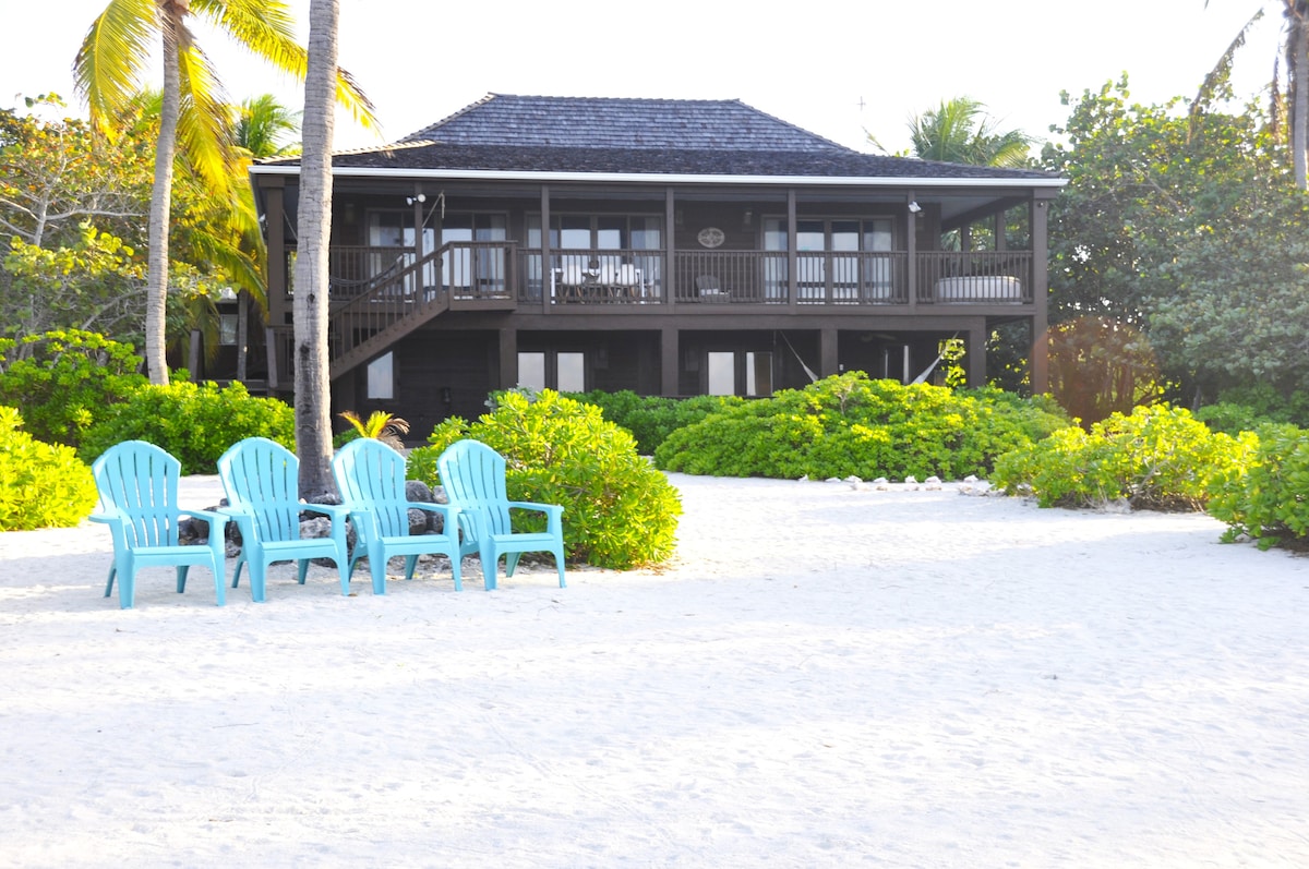 A two-story structure is shown against a backdrop of tropical greenery, featuring multiple balconies and dark wooden accents. In the foreground, four light blue lounge chairs sit on a sandy area, inviting relaxation by the beach.