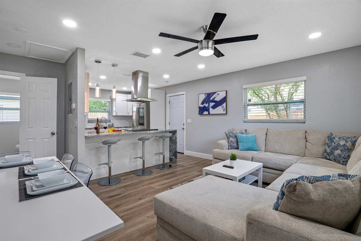 An open living area is presented with a spacious L-shaped couch and a coffee table. A dining area with a table for six is visible alongside the modern kitchen featuring a large island and bar stools. Bright natural light streams through multiple windows.