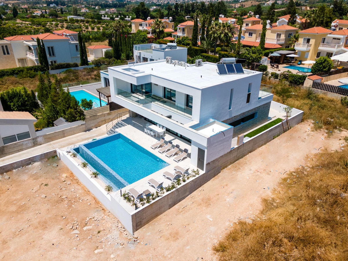 An aerial view captures a modern two-story residence surrounded by lush greenery and neighboring homes. A private swimming pool is featured in the backyard, complemented by sunbeds and landscaped areas for outdoor relaxation. The structure displays a sleek design with large windows and balcony spaces.