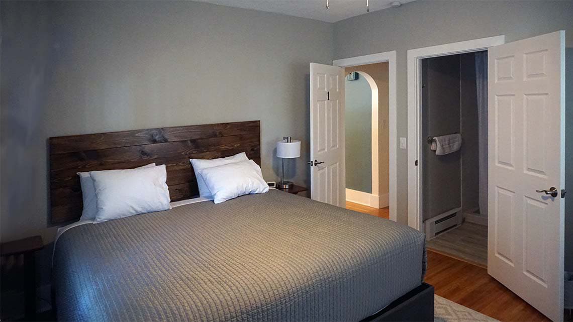 A spacious room is equipped with a comfortable bed featuring a wooden headboard and neutral-toned bedding. Two doors lead to adjacent spaces, including a private full bathroom. A bedside lamp sits on a small table beside the bed, providing soft lighting.
