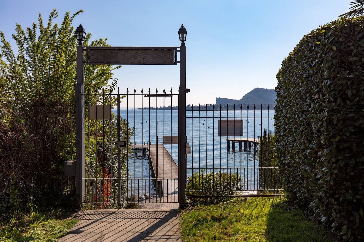 A wrought iron gate opens to reveal a scenic view of the lake, where a wooden pier extends into the water. Lush greenery flanks the pathway leading to the gate, creating a refreshing outdoor space under clear blue skies.
