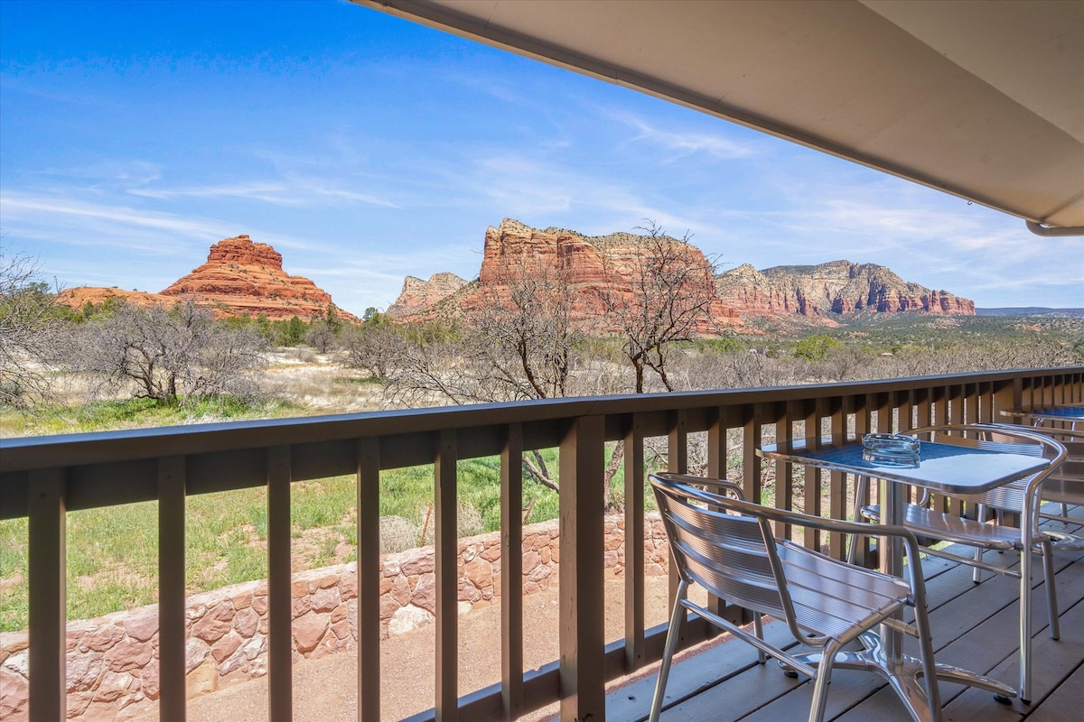 An outdoor patio area is shown, featuring a table and chairs, offering unobstructed views of the red rock formations. The scenery includes vibrant landscape elements, with clear blue skies enhancing the natural beauty of the surroundings.