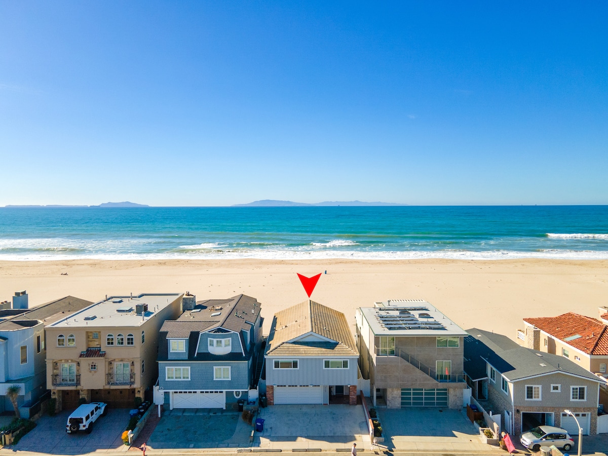 An aerial view of a coastal neighborhood highlights the beachfront location of the property. Waves gently lap the shore, and a clear blue sky stretches overhead. Nearby homes with varied architectural styles line the street, emphasizing the oceanfront setting.