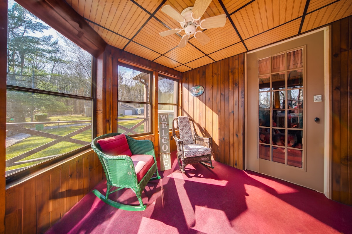 A sunlit sunroom features wooden walls and a plush maroon carpet. Two chairs, one a green rocking chair and the other upholstered with a floral pattern, are arranged to welcome relaxation. A ceiling fan hangs above, and a decorative sign reading 'WELCOME' adds to the inviting ambiance.