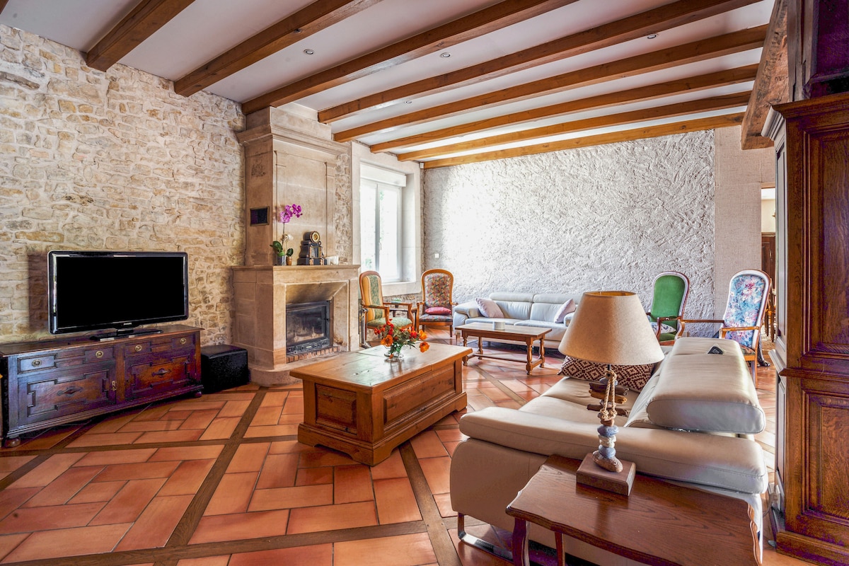 The spacious living room features a stone wall, wooden beams, and terracotta flooring. A fireplace and a television are visible, along with various seating options that include sofas and armchairs. Natural light fills the area through large windows.