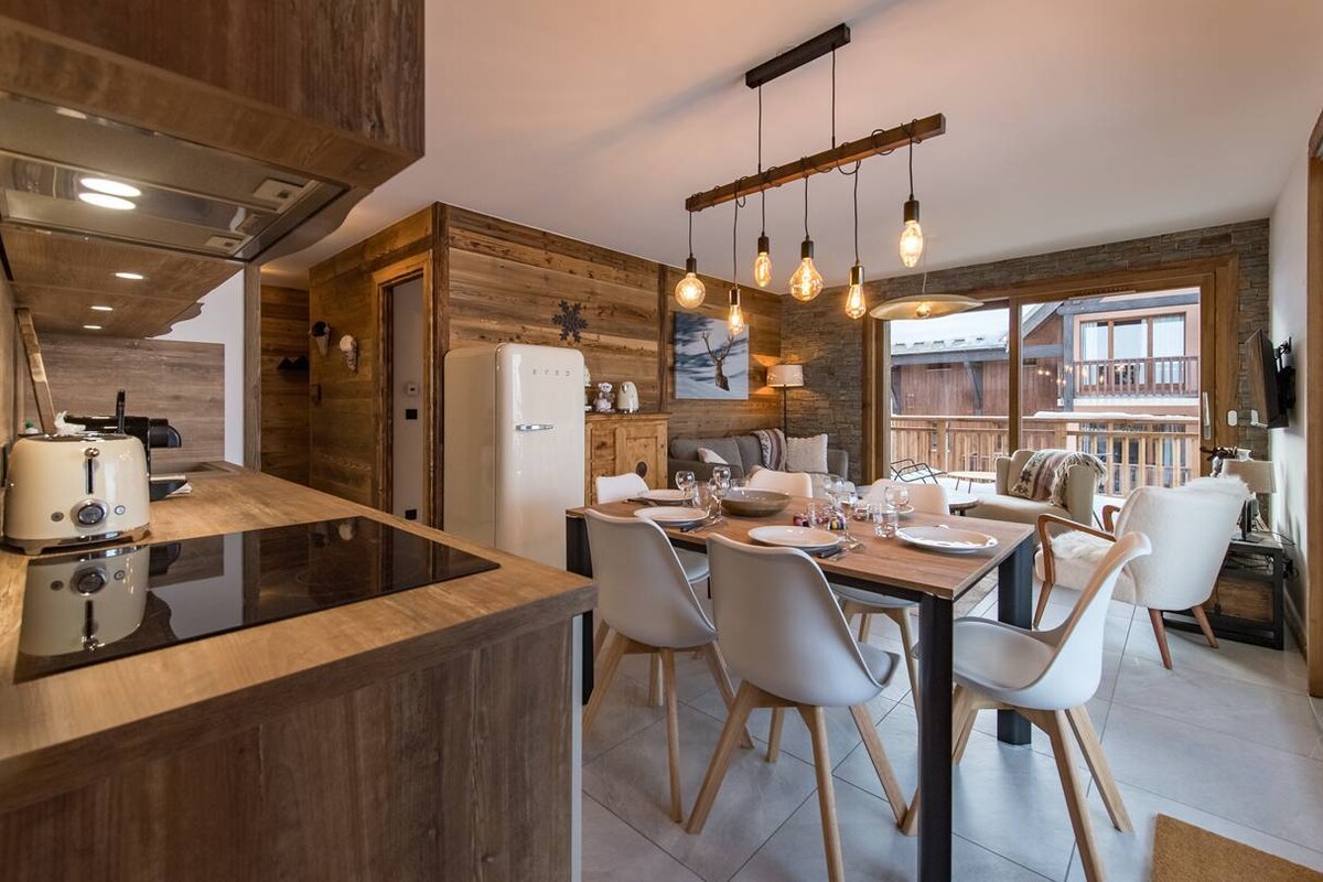 The modern kitchen and dining area features wooden accents and a spacious table set for six. A contemporary refrigerator and induction hob are included, with pendant lighting illuminating the space. Large windows provide natural light and views of the balcony outside.