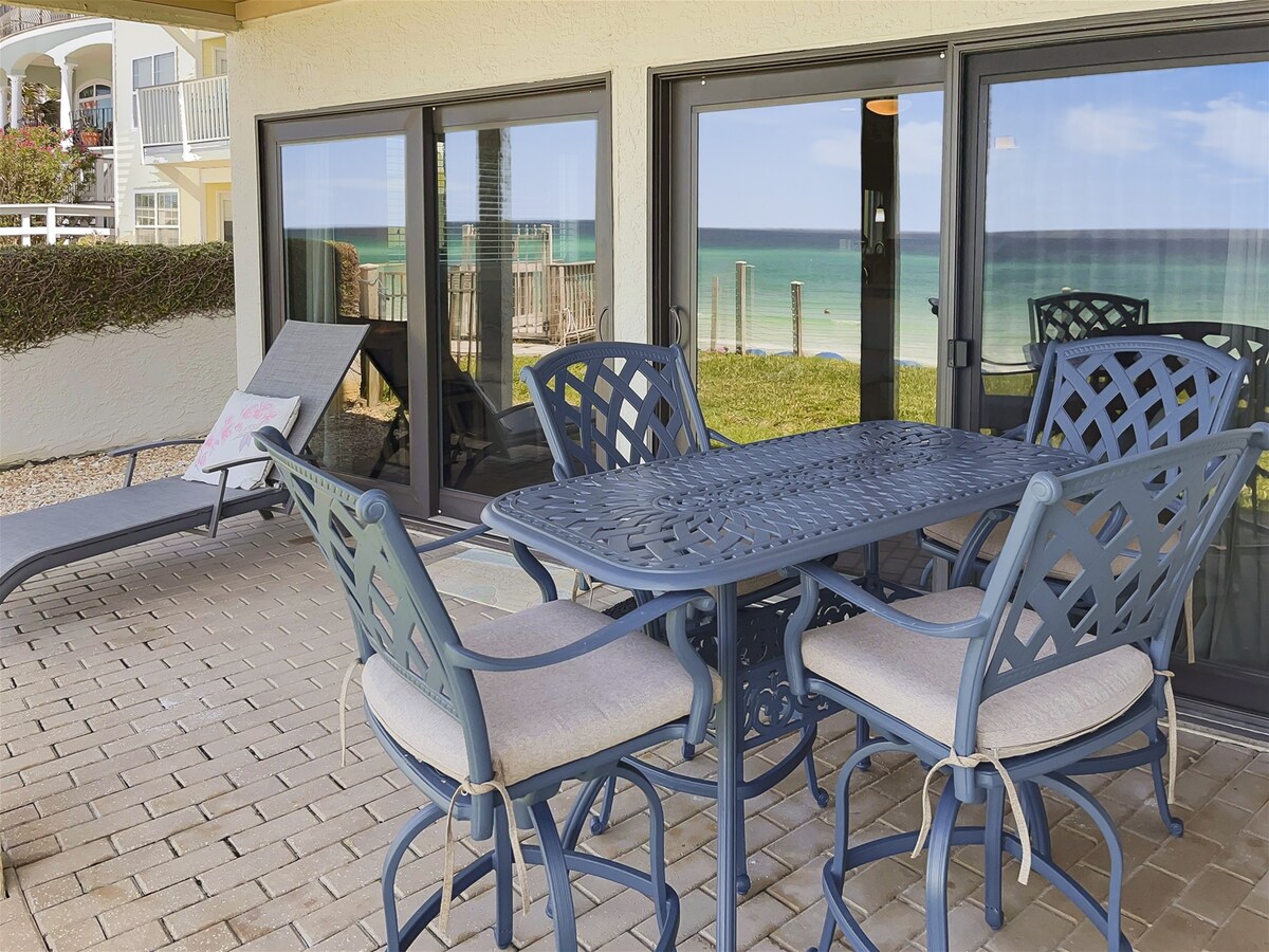 A covered patio area features a dining table with six chairs, complemented by a lounge chair for relaxation. Large sliding glass doors provide panoramic views of the beach and ocean beyond, with the lush green lawn in the foreground.