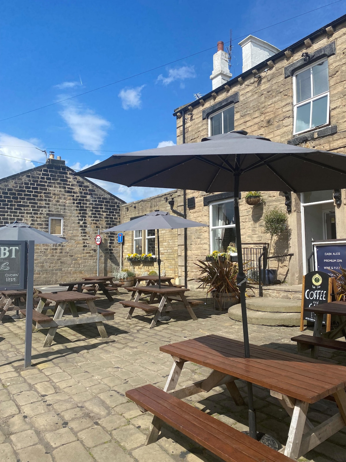 An outdoor seating area features several wooden picnic tables under large grey umbrellas. The space is surrounded by stone walls and greenery, with sunlight filtering through a clear blue sky. A sign indicating 'Coffee' and other offerings is also visible.