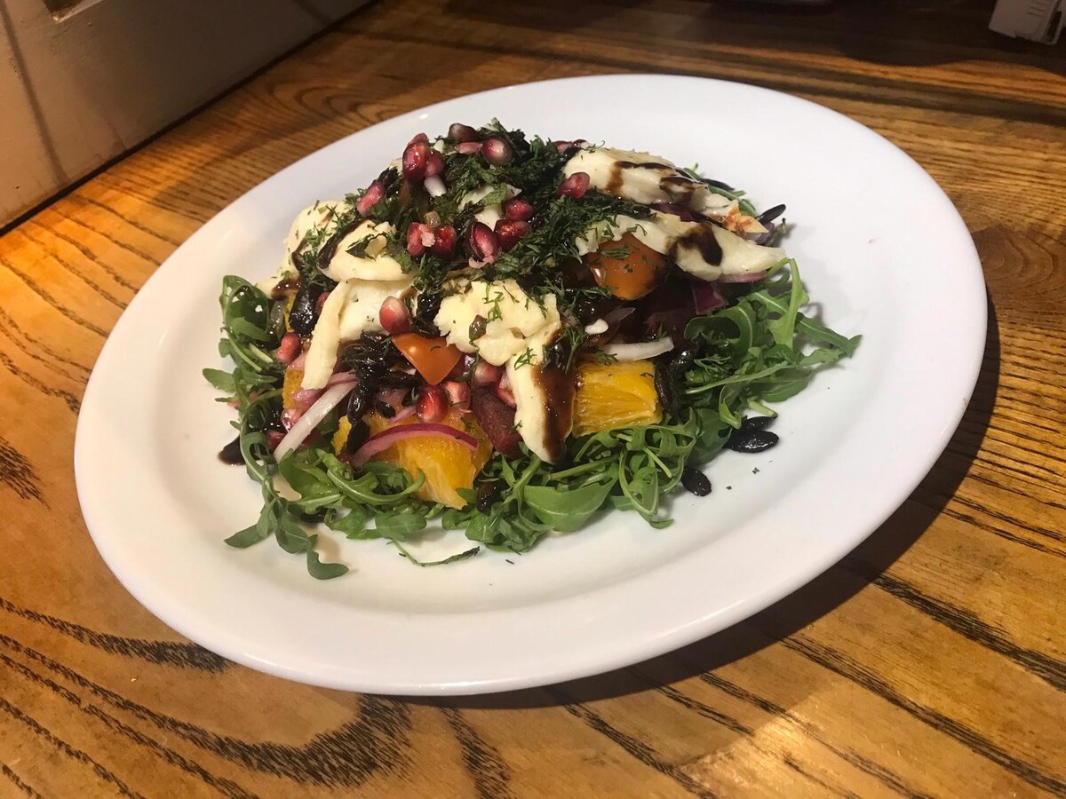 A vibrant salad is presented on a white plate, featuring fresh leafy greens topped with colorful vegetables such as orange slices, tomatoes, and red onion. Pomegranate seeds and herbs are sprinkled on top, while a rich dressing adds depth to the dish.