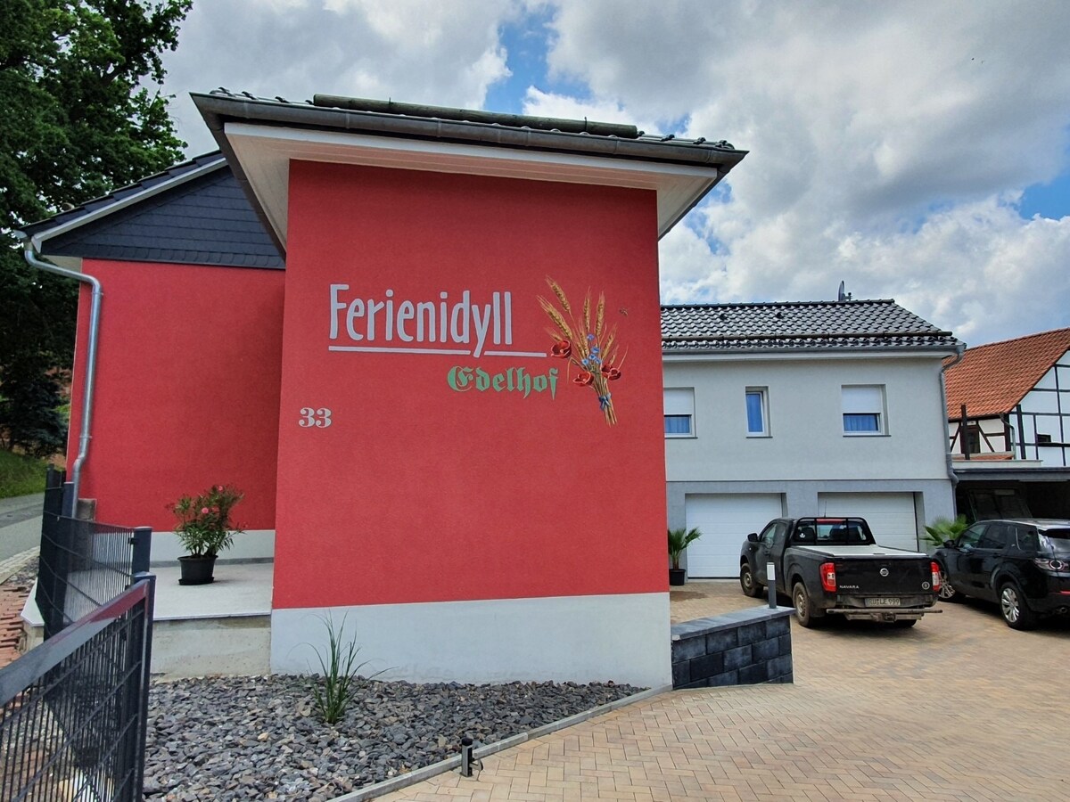 The exterior of Ferienidyll 'Edelhof' is displayed, featuring a vibrant red wall with the property name prominently featured. A paved driveway leads to the building, which includes a garage and ample parking space. Lush greenery surrounds the entrance, enhancing the inviting appearance.