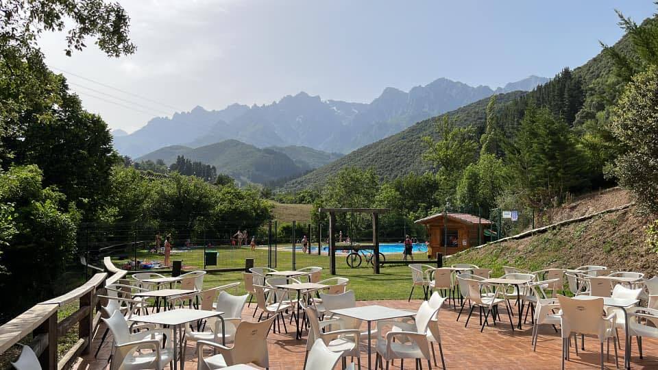 An outdoor seating area is presented with several white chairs surrounding wooden tables, overlooking a grassy space. In the background, a public pool is visible, framed by lush greenery and mountainous terrain, creating a serene atmosphere.