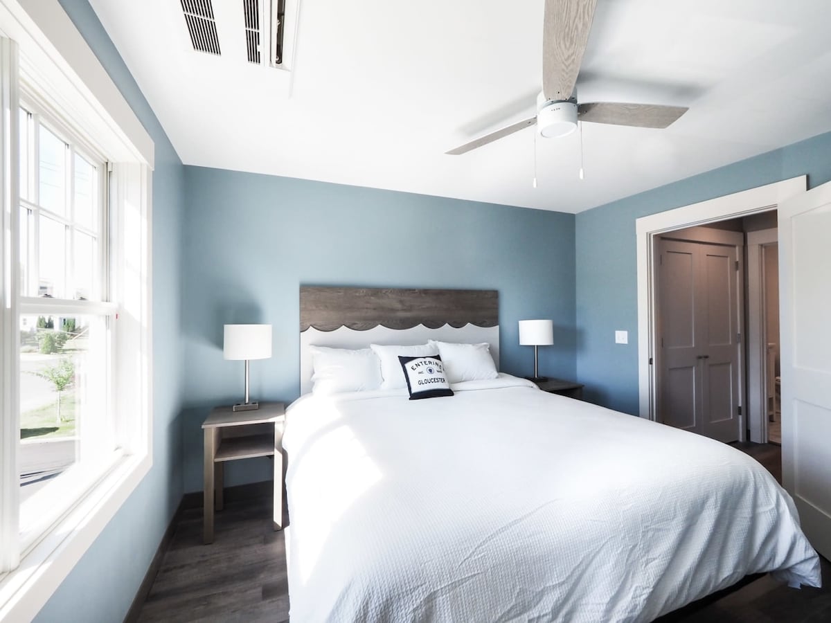 The well-lit bedroom features a spacious bed with crisp white linens and a decorative pillow. Two bedside lamps offer soft lighting, and a ceiling fan provides airflow. A large window with a view allows natural light to illuminate the light blue walls, enhancing the serene atmosphere.