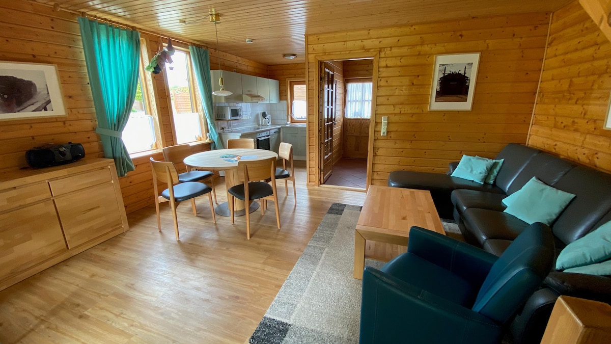 A spacious living area features light wood walls, a comfortable seating arrangement with a dark leather sofa, and a round dining table surrounded by chairs. Large windows, dressed in teal curtains, invite natural light, enhancing the homey atmosphere. A kitchen area is visible in the background.