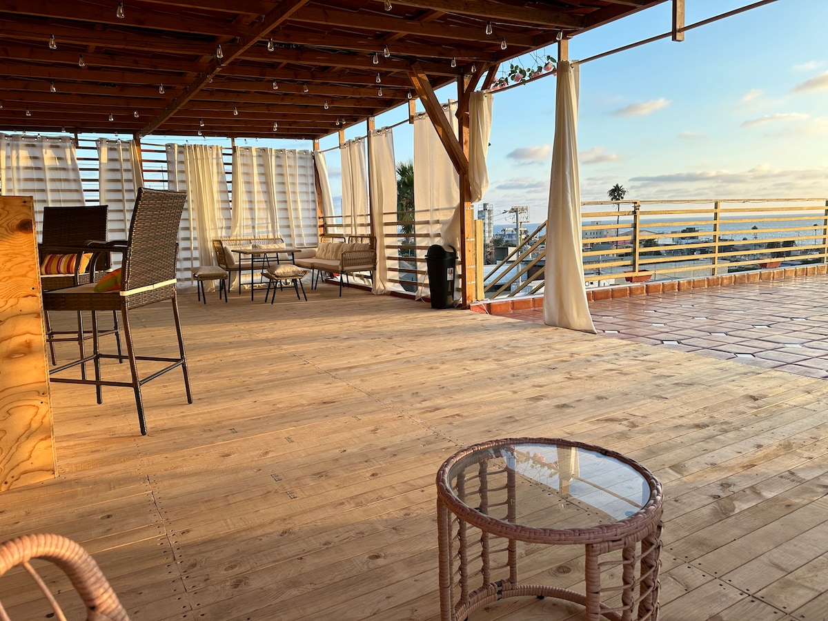 An outdoor terrace features wooden flooring, providing ample space for relaxation and socializing. Weathered furniture includes a dining table and casual seating. Light curtains create a sense of privacy while allowing natural light to filter in, with distant views of the city and beach beyond.