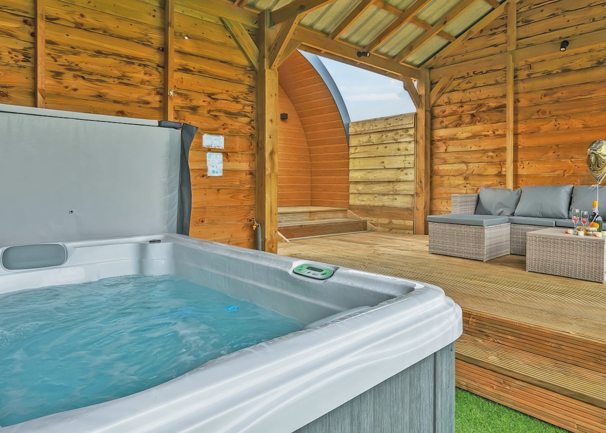 An outdoor hot tub is positioned near a wooden deck area, surrounded by warm-toned wooden walls. A comfortable sofa set, featuring plush cushions, is situated on the deck, offering a relaxed setting for enjoying the space.