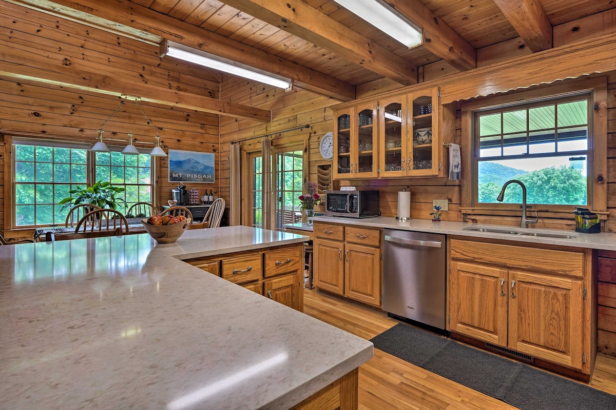 The kitchen area features warm wood cabinetry and a spacious countertop with seating. Stainless steel appliances are visible, including a microwave and dishwasher. Large windows frame views of the surrounding landscape, enhancing natural light in the inviting space.