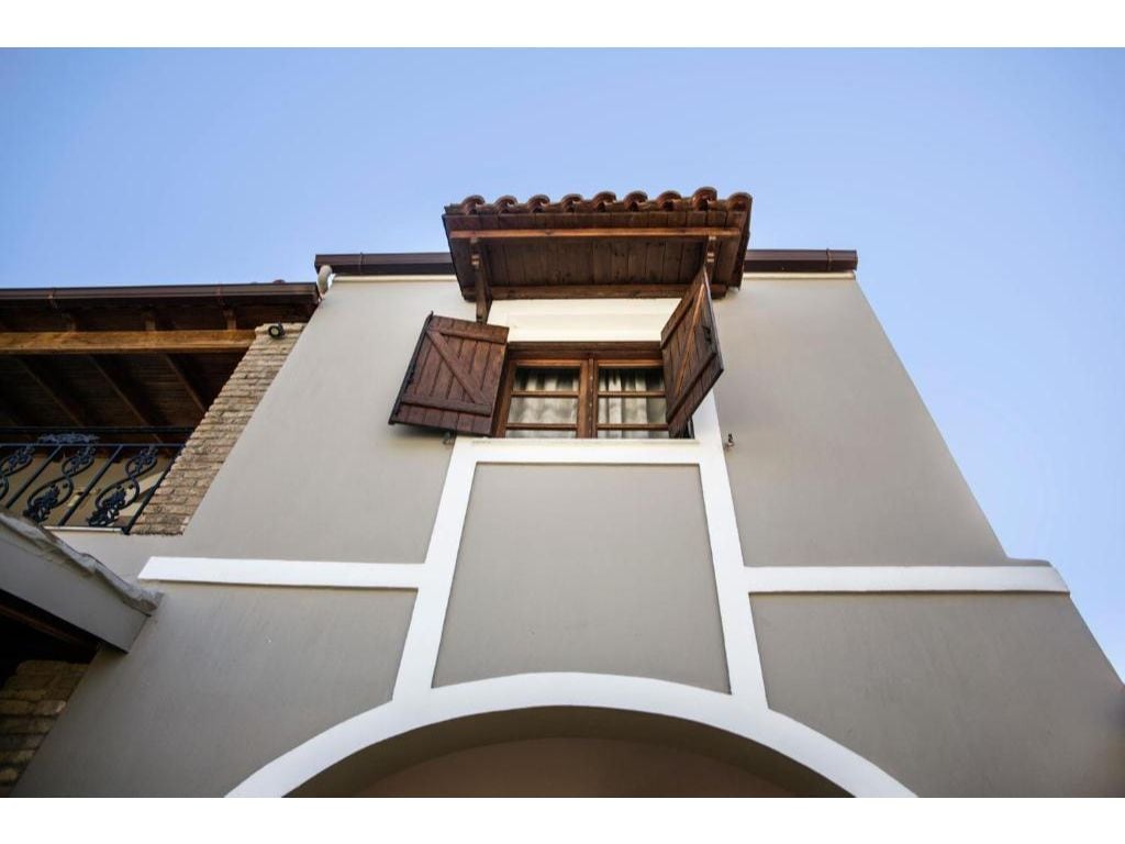 The exterior of the building showcases a charming design with a textured beige wall and decorative elements. A pair of wooden shutters flanks a window, which adds architectural interest. The roof features traditional tiles, enhancing the character of the property against a clear blue sky.