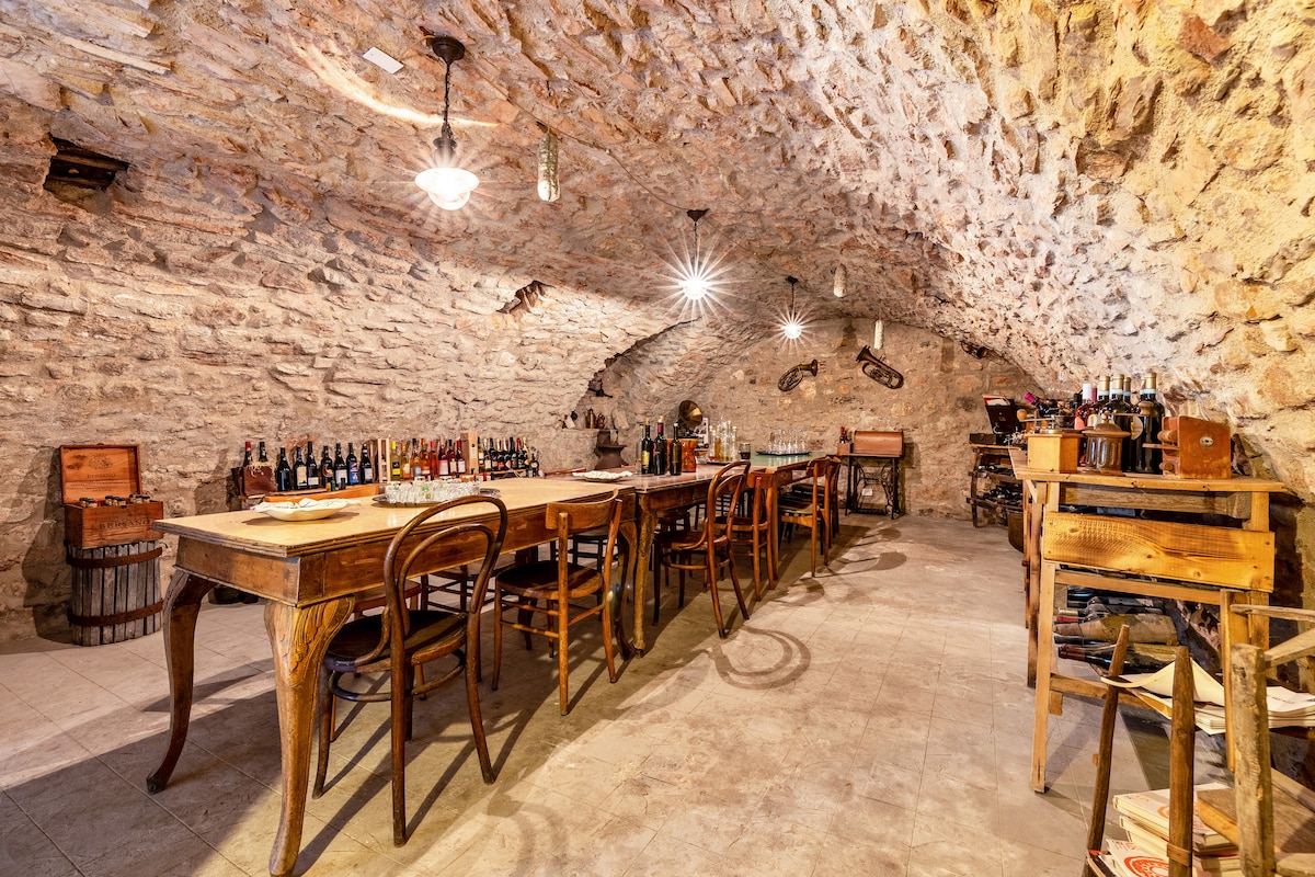 A spacious cellar is highlighted by arched stone walls and a vaulted ceiling. A long wooden table with several wooden chairs is placed centrally, while various bottles of wine are neatly arranged along the walls. Soft lighting creates a warm ambiance throughout the space.
