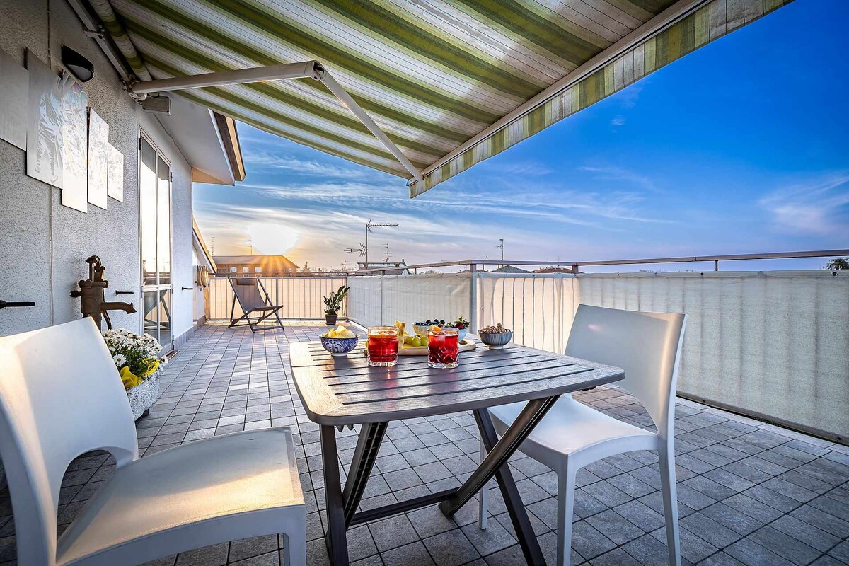 An outdoor terrace features a wooden table set for drinks with two white chairs. A retractable awning provides shade, while a horizon of soft colors is seen in the sky at sunset. Potted plants add greenery to the space.