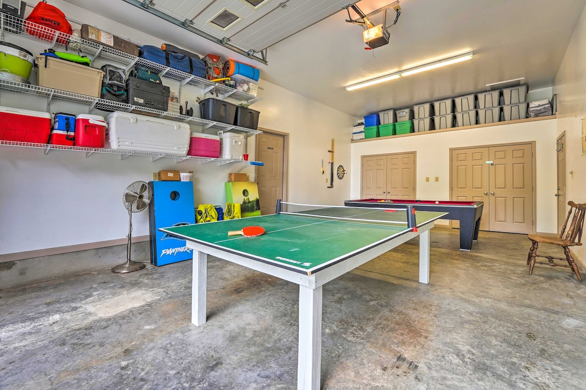 A spacious garage features a ping pong table in the center, surrounded by various storage bins arranged on shelves. A fan stands in one corner, and another table is positioned against the wall, providing additional recreational space.