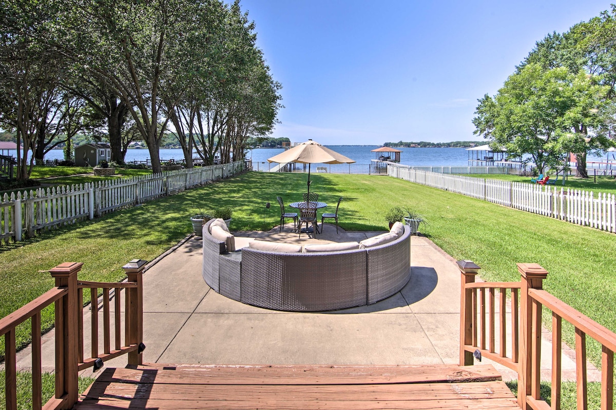 An outdoor entertainment space is shown, featuring a circular seating area with a large umbrella. Lush green grass surrounds the patio, leading to the tranquil lake visible in the distance. A white picket fence borders the yard, enhancing the scenic lakeside view.