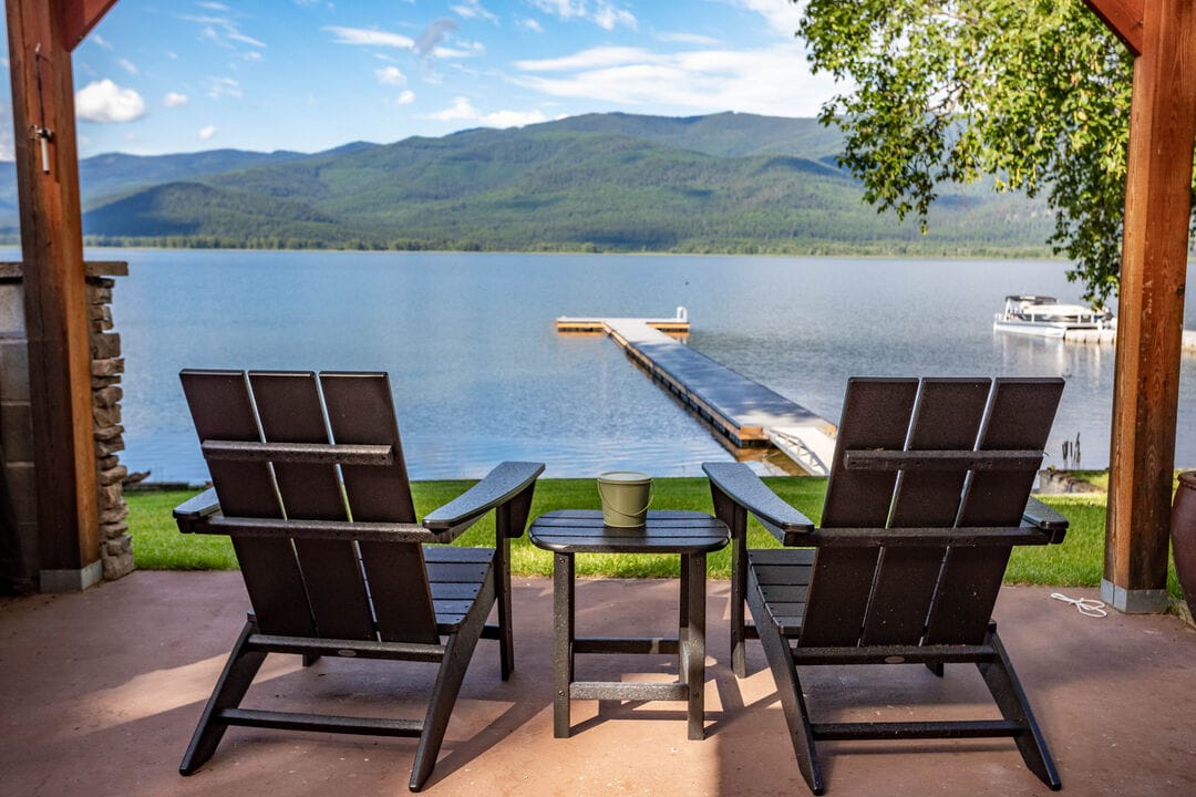 Two comfortable lounge chairs are positioned facing a serene lake, offering unobstructed views of the water and surrounding mountains. A small table is placed between the chairs, enhancing the inviting outdoor space. The dock extends into the lake, providing easy access to water activities.