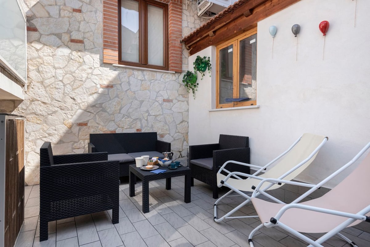 A private terrace features a seating area with dark wicker furniture and a small table, accompanied by two reclining lounge chairs. Sunlight creates soft shadows across the light-colored tiles, while green plants and decorative elements adorn the walls, enhancing the serene outdoor space.