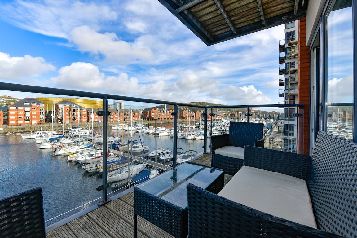 A balcony is shown featuring rattan-style seating and a glass table, overlooking the marina. Boats are moored in the water, and a mix of buildings is visible in the distance under a partly cloudy sky.