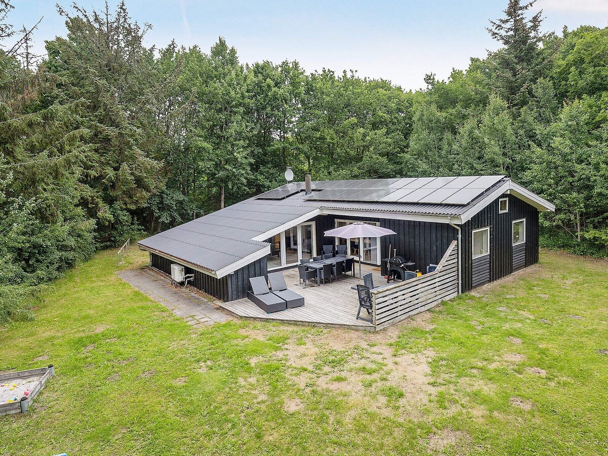 A modern holiday home is set within a green landscape, featuring a large patio area with outdoor furniture and a shaded table. Solar panels are visible on the roof. Surrounding trees provide privacy, enhancing the home's peaceful environment.