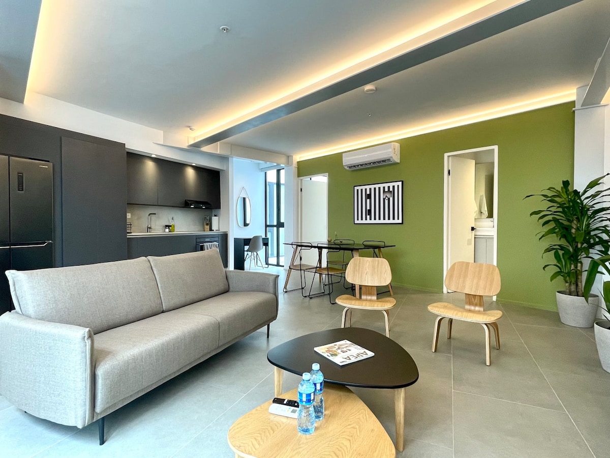 A modern living area is presented with a light grey sofa positioned in front of a sleek black coffee table. Two wooden chairs are set beside the table. An accent wall in vibrant green provides a pop of color, while a doorway leads to another room.
