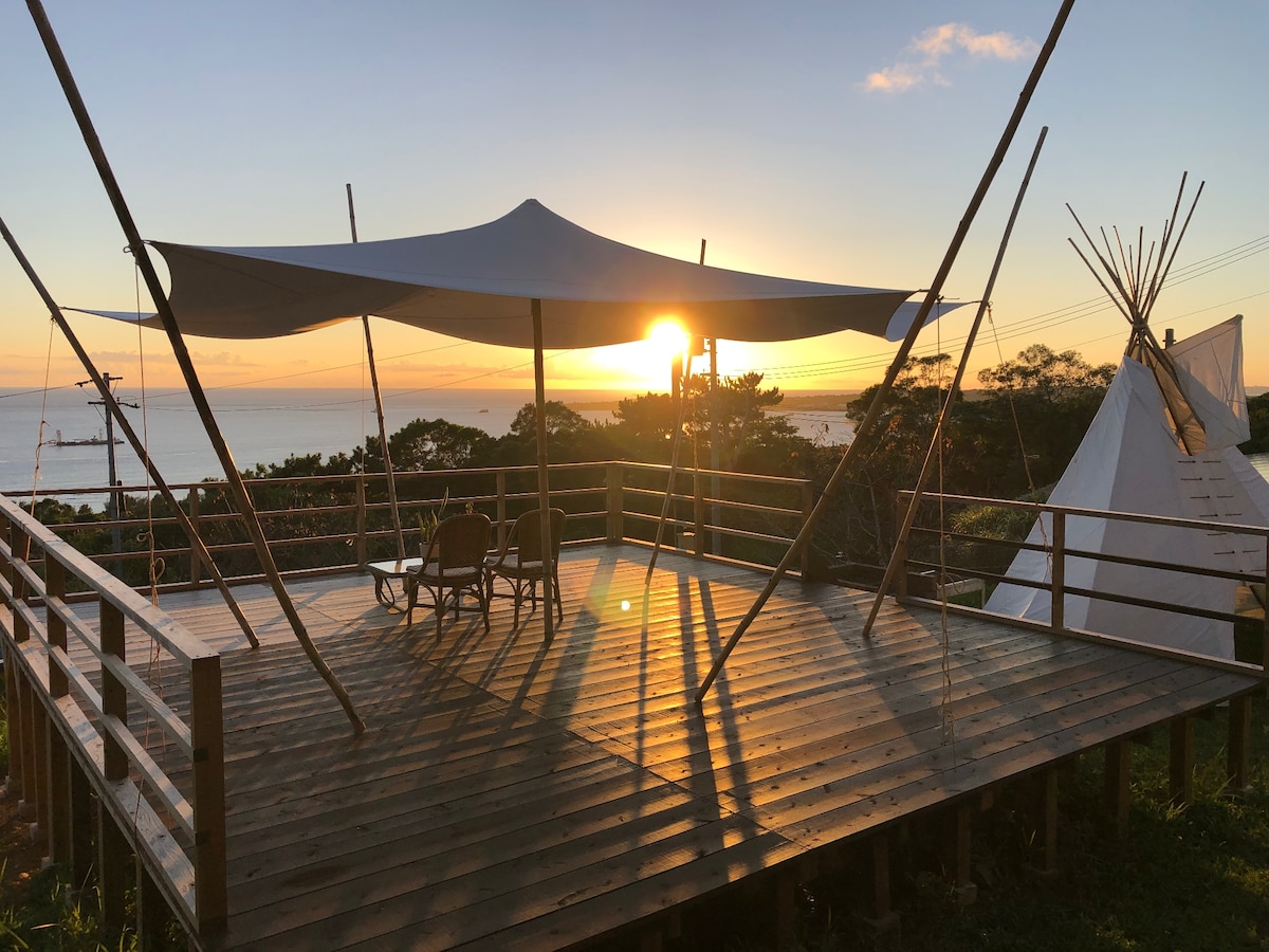 A wooden deck is presented at sunset, featuring a large canopy providing shade. A pair of chairs is positioned to face the ocean view. On one side, a teepee structure is visible against the horizon, where the sun gracefully descends.