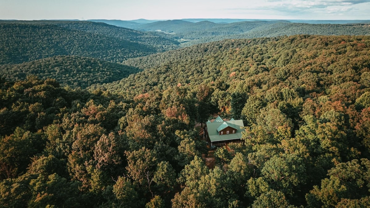 Aerial view showcases a luxurious home nestled amid lush greenery on a vast landscape of rolling hills and valleys. The surrounding 200 acres of private woodland are visible, emphasizing the property's secluded nature and connection to the natural environment.