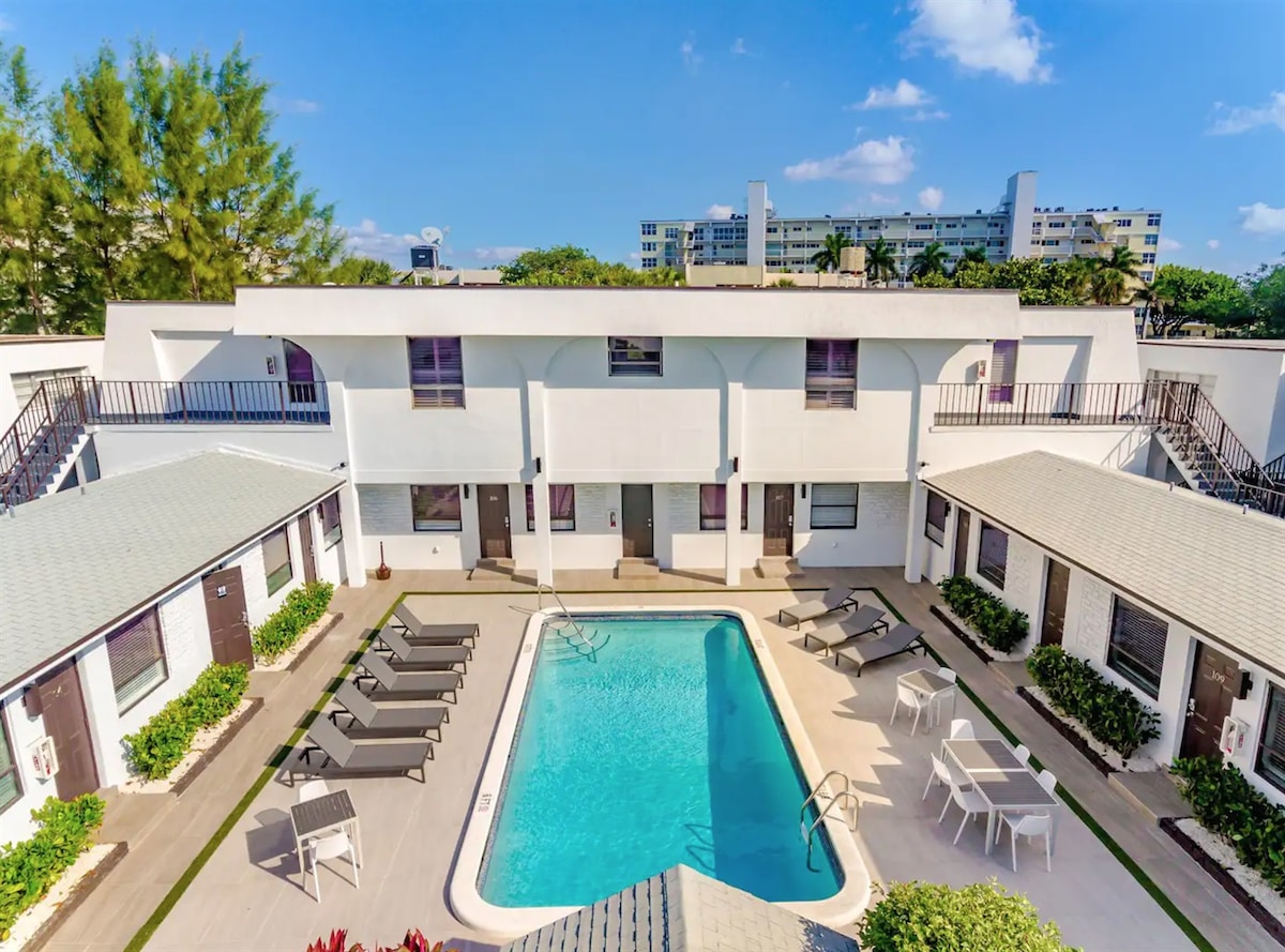 The courtyard features a heated swimming pool surrounded by sun loungers and tables. The two-story structure, painted white, has multiple doorways leading to the apartments. Lush greenery is visible in the background, enhancing the inviting outdoor space.