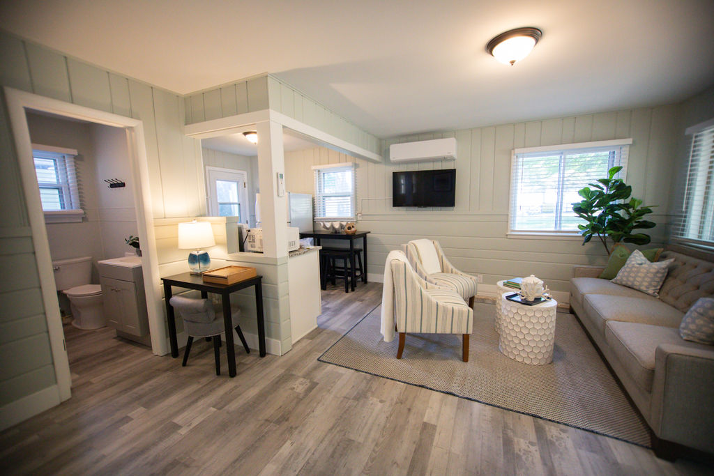 A light-filled living area features a comfortable sofa and two accent chairs, arranged around a textured area rug. A small dining table sits adjacent to the kitchenette, with soft lighting and greenery enhancing the warm atmosphere. Large windows allow natural light to fill the space.