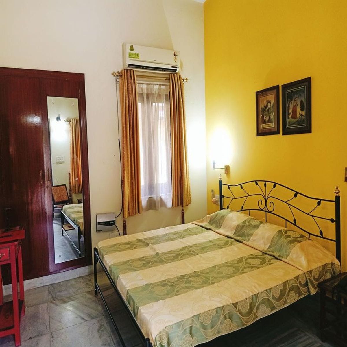 A bright room features a spacious bed with an olive and cream patterned bedspread. Yellow walls enhance the light from the window, adorned with sheer curtains. A wooden cabinet and red side table provide storage, while framed art adds character to the space.