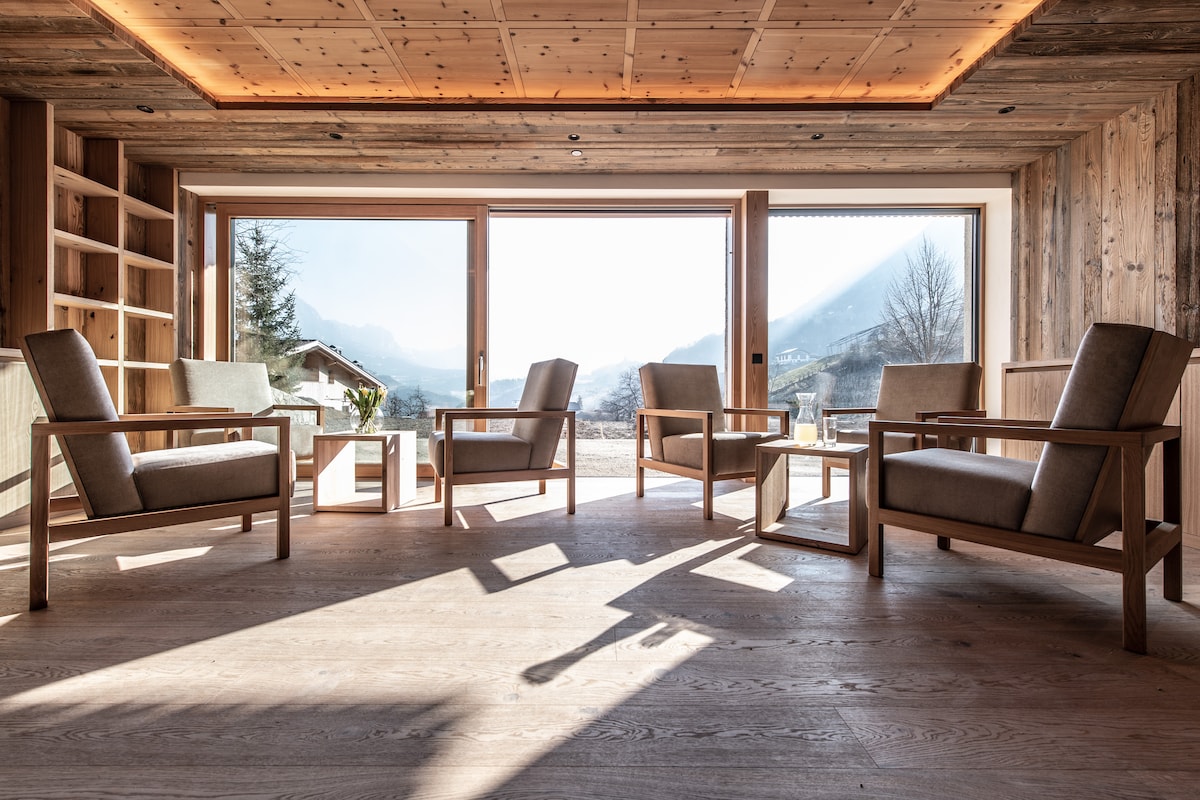 A bright living area features large glass windows, allowing natural light to illuminate the space. Simple wooden chairs are arranged around a small table, with a scenic view of the mountains visible in the background.